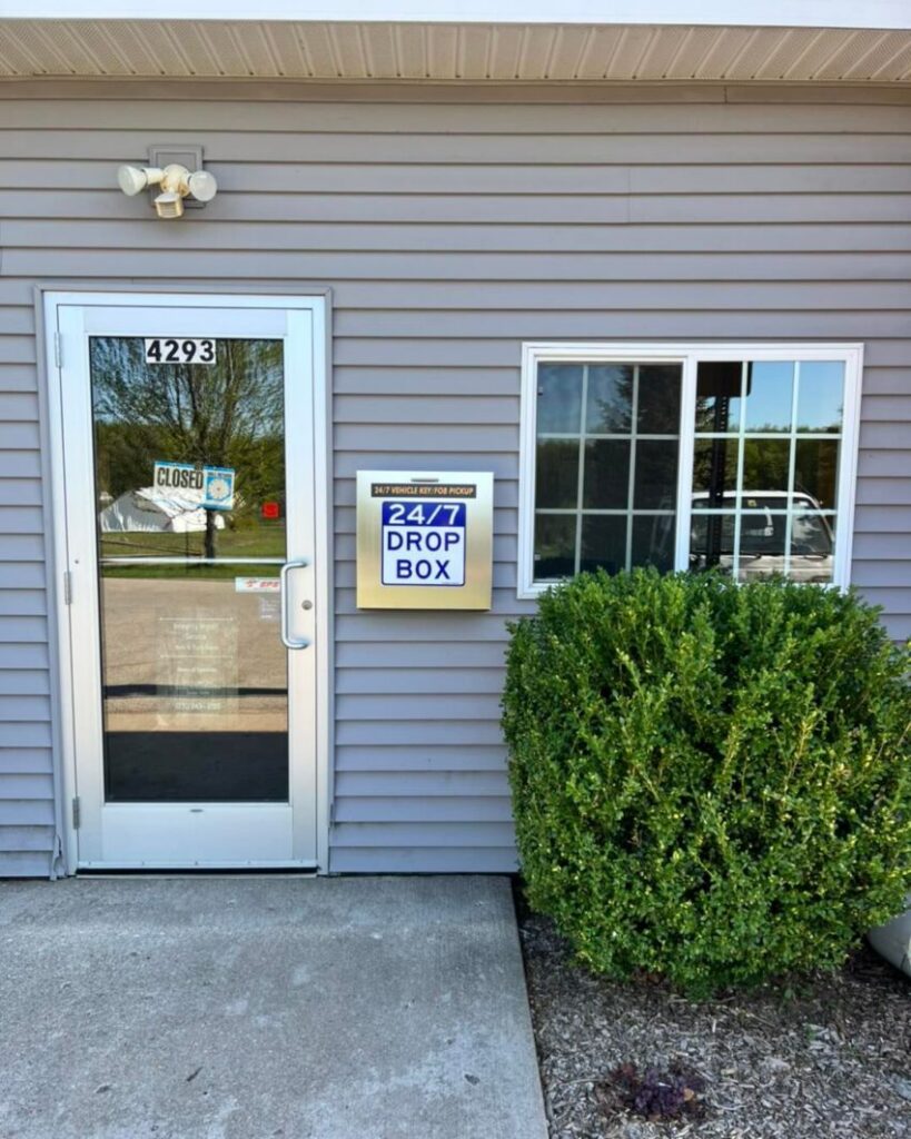 A building entrance with a glass door labeled 4293 and a Closed sign. Next to the door is a wall-mounted box labeled 24/7 Drop Box. A bush and a window are beside the drop box.