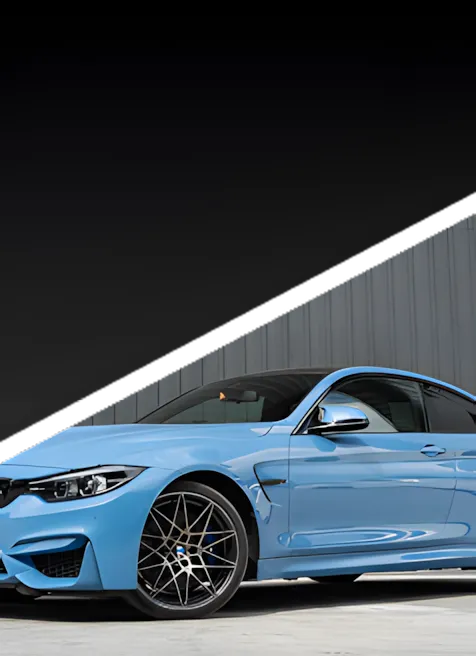 A light blue sports car with black wheels is parked on a concrete surface in front of a gray metal wall, with a dark gradient sky in the background.