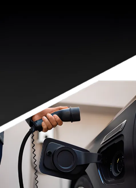 A person’s hand holds a charging plug, preparing to connect it to an electric vehicle’s charging port. The upper part of the image is obscured by a dark, diagonal overlay.
