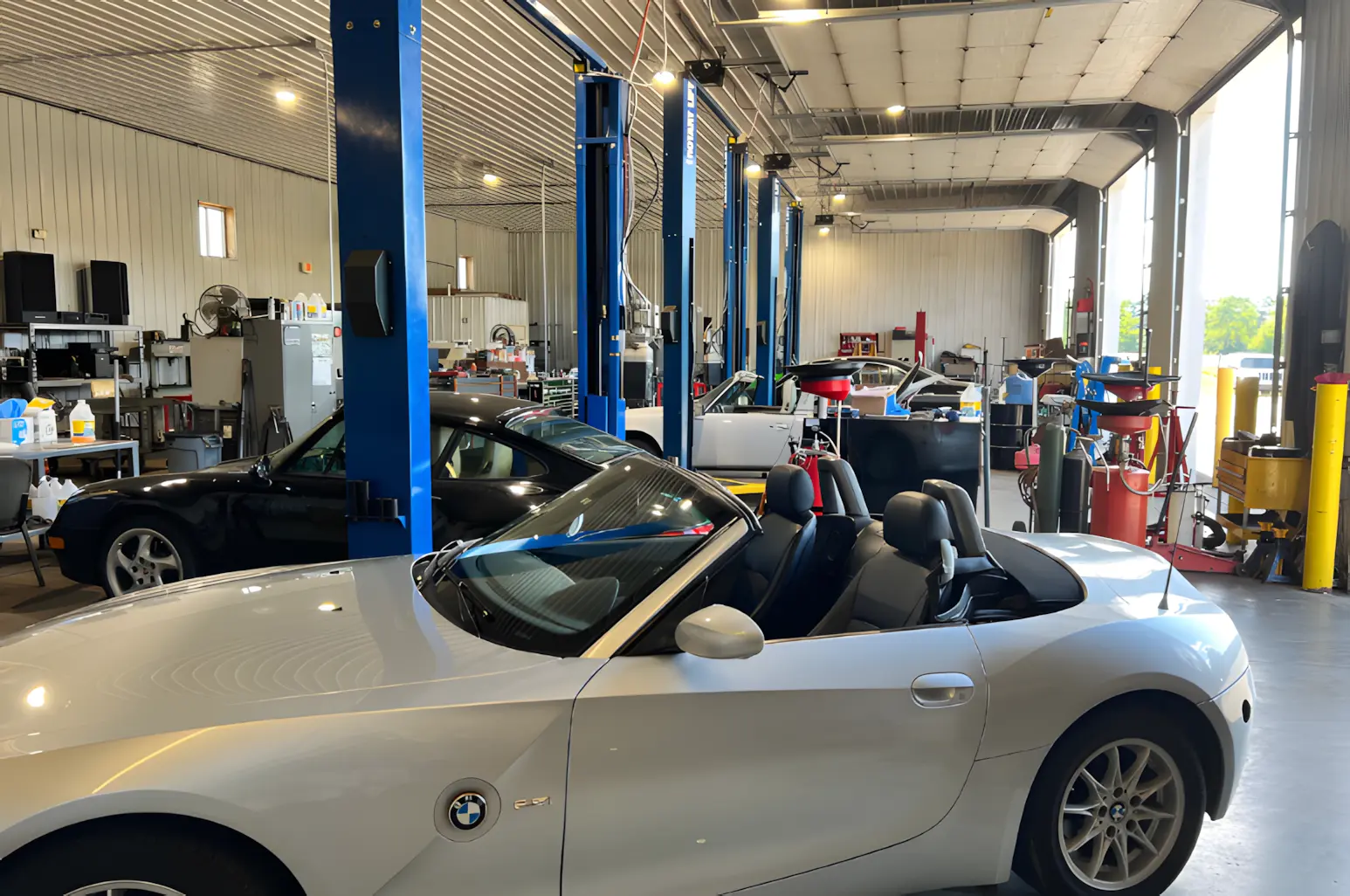A bright, spacious auto repair shop with several cars, including a white BMW convertible in the foreground, and multiple service bays and equipment visible in the background.