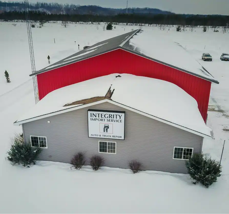 About us, auto repair in Grawn, MI at Integrity Import Service. A building labeled Integrity Import Service Auto & Truck Repair is covered in snow, with part of the roof collapsed. A large red barn is visible behind it, and the ground around is snowy with some scattered trees.