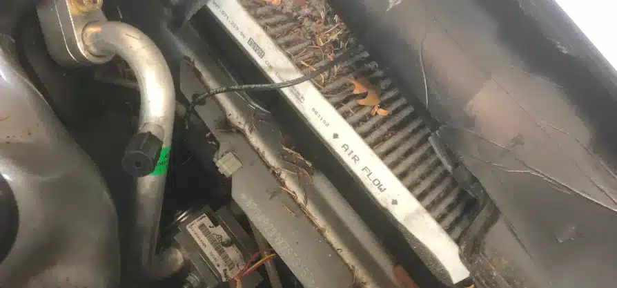 Auto a/c and heater repair in Grawn, MI at Integrity Import Service. A dirty car cabin air filter covered in dust, leaves, and debris, partially removed from its housing under the hood. The words AIR FLOW are visible on the filters edge.