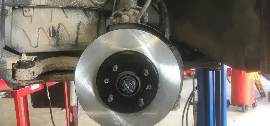 Brake repair in Grawn, MI at Integrity Import Service. A close-up view of a cars wheel hub showing a new, shiny brake rotor installed, with part of the suspension and undercarriage visible in a workshop setting.