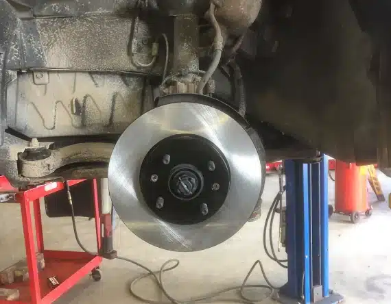 A close-up view of a shiny new brake disc installed on a car wheel hub, with the vehicle lifted on a hydraulic lift in an auto repair shop. Various tools and equipment are visible in the background.