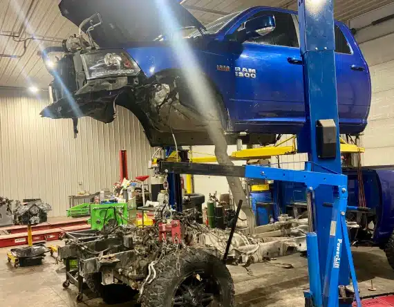 A blue RAM 1500 pickup truck is elevated on a hydraulic lift in an auto repair shop, with the front end removed and the chassis and engine separated beneath it. Various tools and equipment are visible around the workspace.