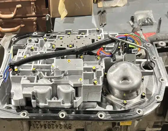 Open transmission valve body with exposed wiring, connectors, and internal metal components, set on a workshop table surrounded by tools and parts in the background.