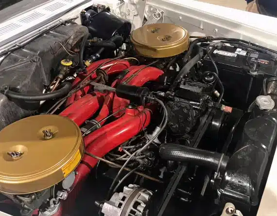 Close-up view of a car engine compartment featuring two gold air filter covers, red intake manifolds, and various black components, wiring, and a car battery on the right side.