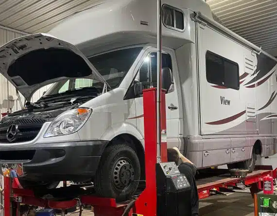 A white recreational vehicle (RV) with a View logo is lifted on a red hydraulic car lift in a garage. The RVs front hood is open, and a mechanic is working underneath it.