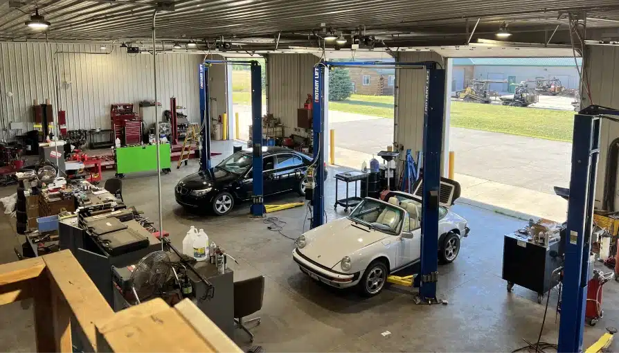 Vehicles we service, auto repair in Grawn, MI at Integrity Import Service. A spacious automotive repair shop with two cars, one black and one white convertible, parked on lifts. Equipment, tools, and workbenches line the walls; large open doors reveal grass and outdoor machinery.
