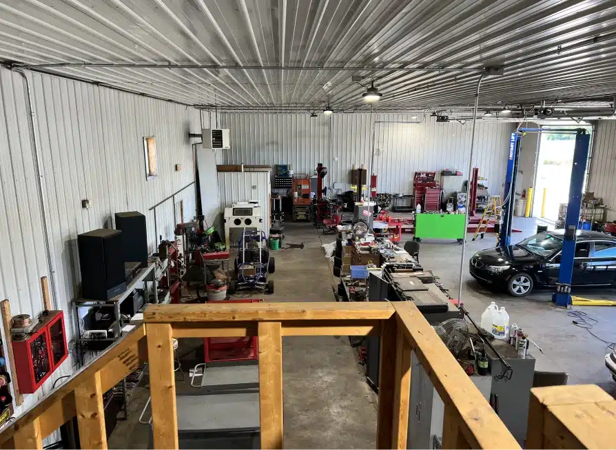 Interior view of a spacious auto repair shop with various tools, workbenches, equipment, and a car on a lift. The shop has a high metal ceiling, white walls, and a concrete floor, with items neatly organized throughout the space.