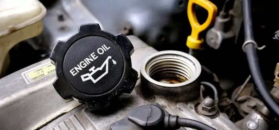 Close-up of an open engine oil cap labeled ENGINE OIL on a car engine, with the oil fill port exposed and a yellow dipstick handle visible in the background.