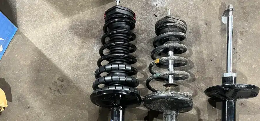 Steering and suspension repair in Grawn, MI at Integrity Import Service. Three car suspension struts are lined up on a concrete floor; the left one is new and shiny, the middle one is old and rusted, and the right one is a new, partially assembled strut.