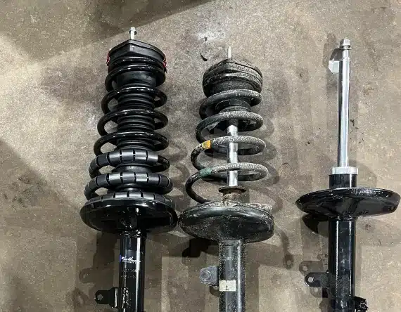Three car suspension struts are laid side by side on a concrete floor: the left one is new and fully assembled, the middle one is old and worn, and the right one is a new strut without a spring.
