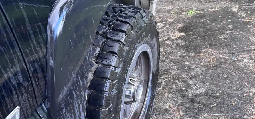 Tire sales & services in Grawn, MI at Integrity Import Service. Close-up of a black off-road vehicle tire with deep treads, mounted on a silver wheel, next to a dark, wet, and slightly muddy concrete surface.