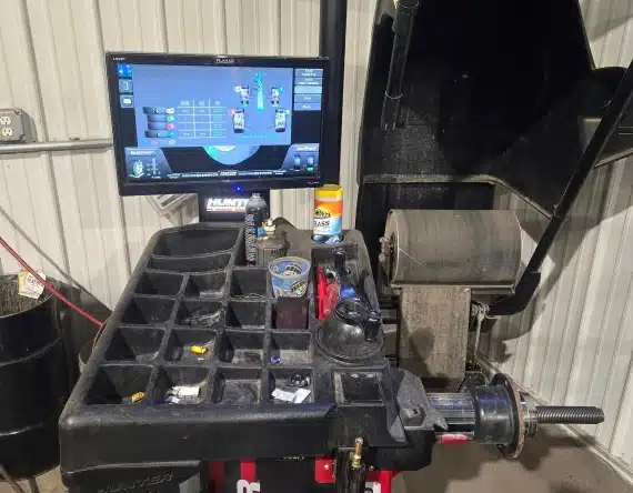 A tire balancing machine with a black organizer tray holding various tools and items, a monitor displaying a tire balancing interface, and part of a wheel balancer visible in a workshop setting.