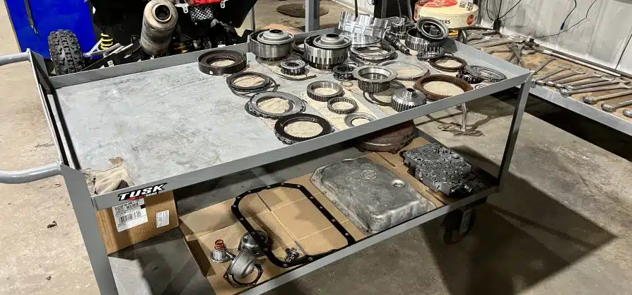 Transmission service & repair in Grawn, MI at Integrity Import Service. A metal cart holds disassembled transmission parts, including gears, gaskets, and seals, neatly organized on the top shelf, with additional automotive components and tools placed on the lower shelf.