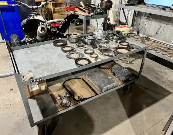 A metal workbench holds assorted gears, bearings, and engine parts, with tools and a cardboard box on the lower shelf. In the background, a small off-road motorcycle is visible in a garage setting.