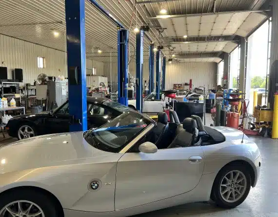 Pre-purchase inspection in Grawn, MI at Integrity Import Service. A white BMW convertible is parked inside an auto repair shop with several work bays, car lifts, tools, and equipment visible in the background. Other cars are also being serviced.