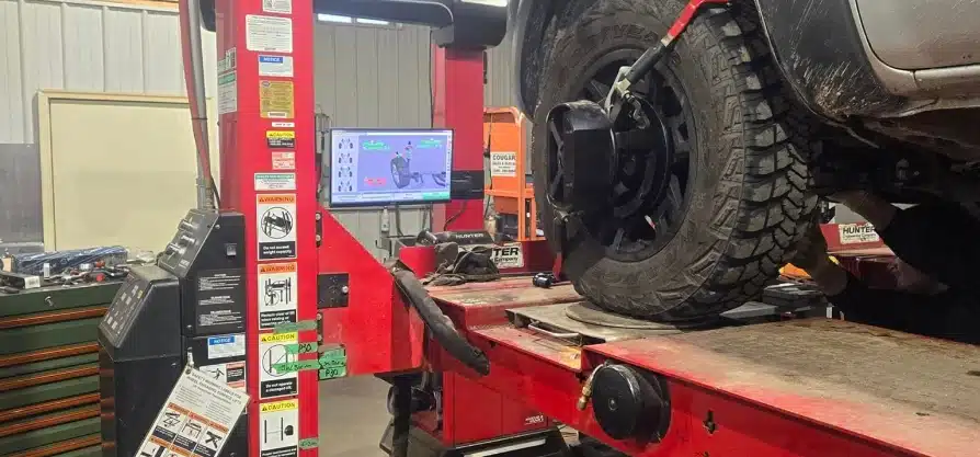 Wheel alignment in Grawn, MI at Integrity Import Service. A vehicle is lifted on a red alignment rack in an auto repair shop, with a wheel alignment machine attached to the tire and a computer screen displaying alignment data in the background.