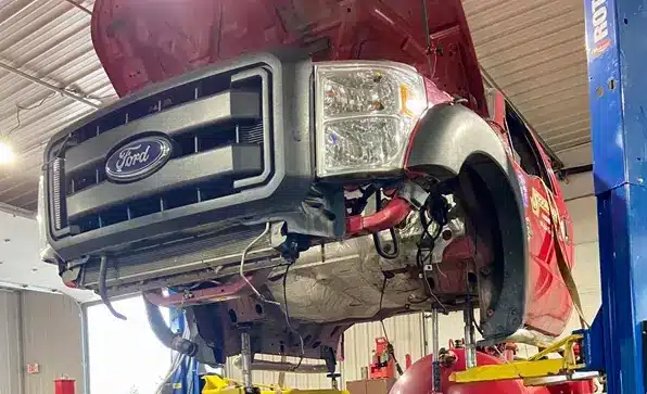Auto and light truck repair in Grawn, MI at Integrity Import Service. A red Ford truck with its hood open is elevated on a hydraulic lift inside a garage, exposing the engine and undercarriage for maintenance or repair.