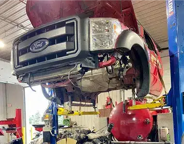 A red Ford truck with its hood open is lifted on a hydraulic car lift inside a garage, exposing its undercarriage for repair or maintenance. Tools and equipment are visible in the background.