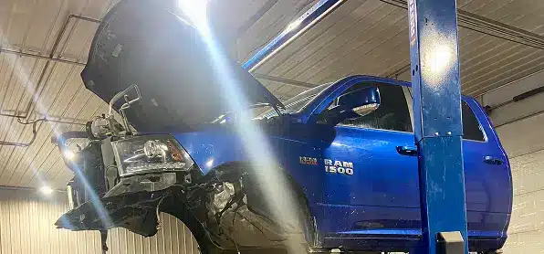 A blue Dodge Ram 1500 truck is lifted on a hydraulic car lift inside an auto repair shop, with its front hood open and front bumper removed. Bright lights illuminate the workspace.