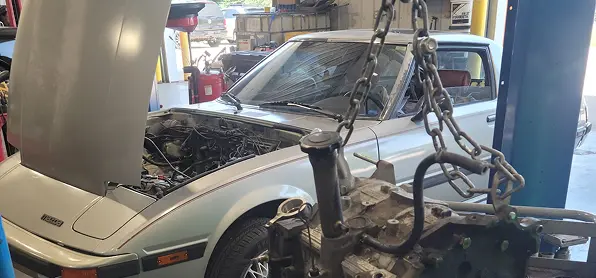 Drivetrain service in Grawn, MI at Integrity Import Service. A silver classic car with its hood open is parked in a garage, with an engine suspended by a chain hoist in the foreground, indicating an engine removal or replacement in progress.