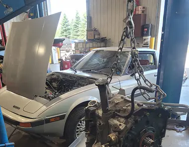 A silver car with its hood open is parked in a garage. An engine is suspended by a chain hoist in the foreground, indicating that engine work or replacement is being performed. Tools and equipment are visible around the workspace.