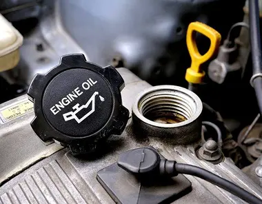 Close-up of a car engine with the oil cap removed. The cap, labeled ENGINE OIL with an oil can icon, is placed beside the open oil filler port. The oil dipstick is visible in the background.