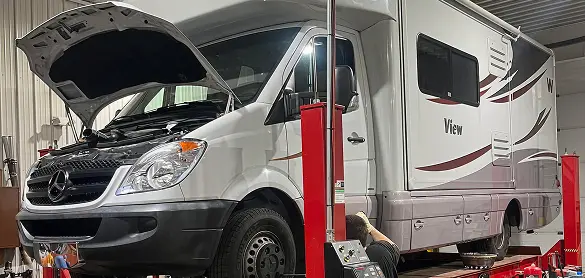 Fleet service in Grawn, MI at Integrity Import Service. A white recreational vehicle (RV) with a View logo is lifted on a red hydraulic car lift in a garage. The RVs front hood is open, and a mechanic is working underneath it.