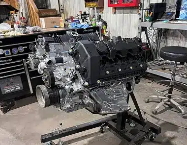 A car engine mounted on a stand in a workshop, surrounded by tools, equipment, and workbenches, with various automotive parts and supplies visible in the background.